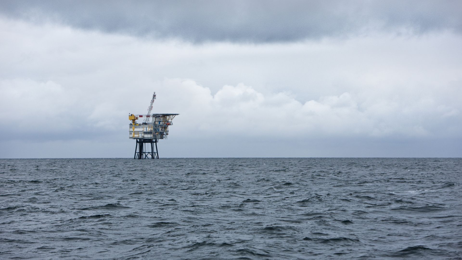 A wide shot of an offshore oil platform, standing on its black legs, in a grey, choppy North Sea. The sky is filled with thick, grey clouds.