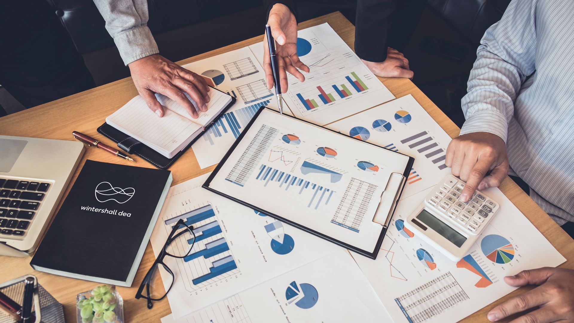 diverse Papiere mit verschiedenen Diagrammen in Blautönen liegen auf einem Tisch neben einem Wintershall Dea Notizbuch. Am Tisch sitzen und stehen drei Personen und scheinen zu diskutieren.